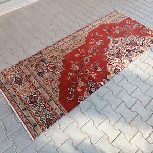 Puede incluir: Una alfombra oriental rectangular roja con intrincados diseños florales en beige, azul y marrón. La alfombra presenta un campo central rojo con un diseño de borde detallado. La alfombra está colocada sobre una superficie de baldosas grises.