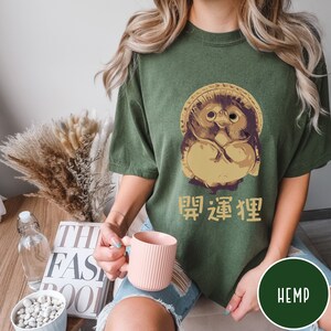 K&ouml;nnte beinhalten: Ein gr&uuml;nes T-Shirt mit einer Comicfigur aus beige und braunem Fell, die einen Hut tr&auml;gt. Die Figur h&auml;lt ihre H&auml;nde vor der Brust zusammen. Der Text "HEMP" befindet sich in der rechten unteren Ecke des Shirts. Der Text "昭 凛 狸" ist auf das Shirt gedruckt.