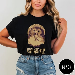 K&ouml;nnte beinhalten: Schwarzes T-Shirt mit einem Cartoon-Bild eines tan- und braunen japanischen Maneki-Neko, oder "Gl&uuml;ckskatze", das einen lila-wei&szlig;en Hut tr&auml;gt. Der Text "開運招福" ist in Gold unter dem Bild gedruckt.
