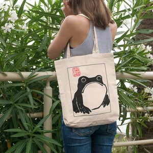 May include: A beige tote bag featuring a black and white illustration of a frog on the front. The frog is looking upwards with a surprised expression. The bag has a red stamp with Japanese characters on it.