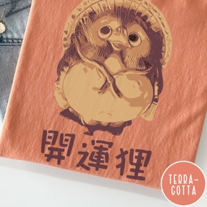 K&ouml;nnte beinhalten: Ein terracottafarbenes T-Shirt mit einem Cartoon-Bild eines japanischen Schutzhundes, bekannt als "Daruma", mit dem Text "開運招福" unter dem Bild.
