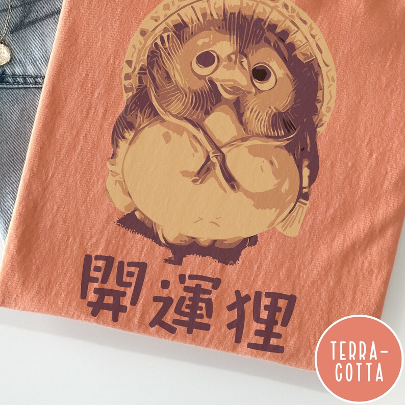 K&ouml;nnte beinhalten: Ein terracottafarbenes T-Shirt mit einem Cartoon-Bild eines japanischen Schutzhundes, bekannt als "Daruma", mit dem Text "開運招福" unter dem Bild.