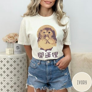 K&ouml;nnte beinhalten: Ein wei&szlig;es T-Shirt mit einem Cartoon-Bild einer japanischen Schutzgottheit, Jizo, mit dem Text "開運狸" in Japanisch. Das Shirt ist in einem hellen Elfenbeinton gehalten.