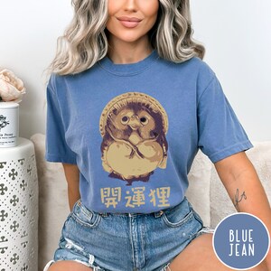 K&ouml;nnte beinhalten: Ein T-Shirt aus Jeansblau mit einem Cartoon-Bild einer japanischen Daruma-Puppe mit Hut. Der Text "開運招福" ist unter dem Bild aufgedruckt.