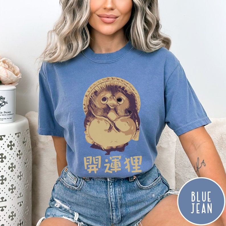 K&ouml;nnte beinhalten: Ein T-Shirt aus Jeansblau mit einem Cartoon-Bild einer japanischen Daruma-Puppe mit Hut. Der Text "開運招福" ist unter dem Bild aufgedruckt.