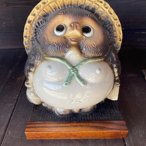K&ouml;nnte beinhalten: Eine Keramik-Tanuki-Figur mit einem braunen und wei&szlig;en Gesicht, gro&szlig;en Augen und einem gr&uuml;nen Band um den Hals. Der Tanuki tr&auml;gt einen Strohhut und sitzt auf einem Holzsockel.