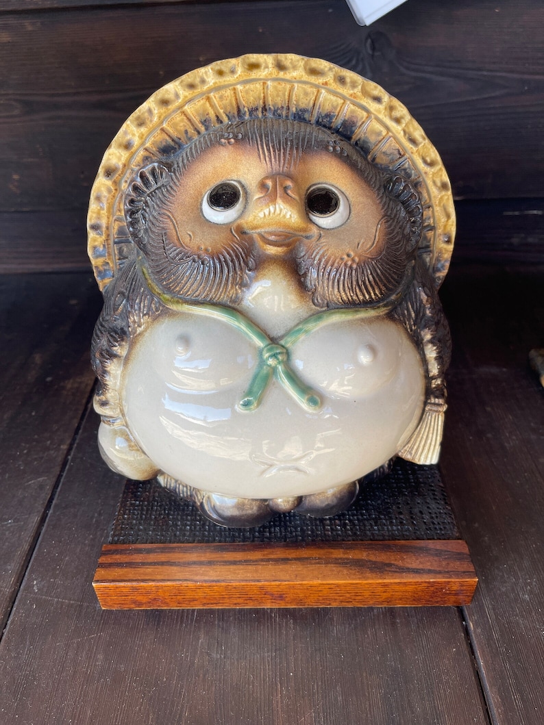 K&ouml;nnte beinhalten: Eine Keramik-Tanuki-Figur mit einem braunen und wei&szlig;en Gesicht, gro&szlig;en Augen und einem gr&uuml;nen Band um den Hals. Der Tanuki tr&auml;gt einen Strohhut und sitzt auf einem Holzsockel.