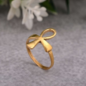 May include: A gold-coloured ring featuring an Ankh symbol. The ring is made of metal and has a smooth finish.
