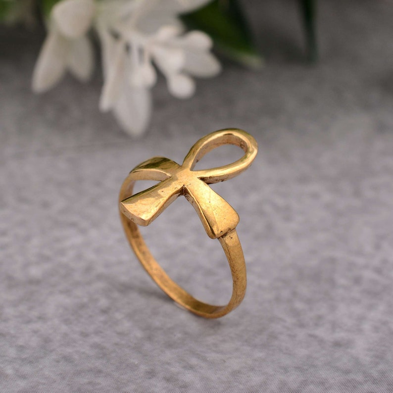 May include: A gold-coloured ring featuring an Ankh symbol. The ring is made of metal and has a smooth finish.