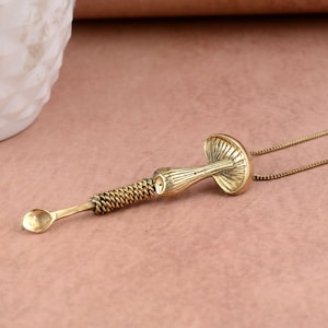 May include: A brass-toned mushroom pendant necklace. The pendant has a small spoon, a coiled section, and a mushroom cap design. The necklace is on a fine chain, and the background is a plain, muted pink.