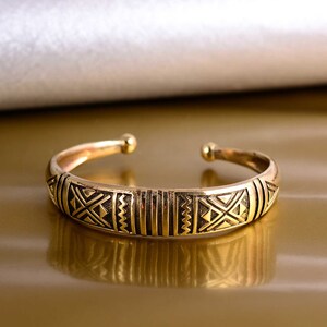 May include: A gold-toned bangle bracelet with a geometric pattern. The bracelet is made of metal and has a simple, elegant design.