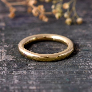 Op de afbeelding: Een goudkleurige ring met een glad, afgerond ontwerp. De ring is cirkelvormig en lijkt van metaal te zijn. De ring staat tegen een donkere, getextureerde achtergrond, met een wazig bloemelement op de achtergrond.