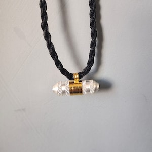 May include: A silver and gold pendant with a black braided cord necklace. The pendant is cylindrical and has a textured surface.