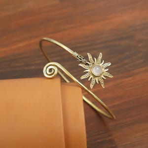 May include: A gold-toned metal cuff bracelet featuring a sun-shaped charm with a central round iridescent stone. One end of the open band terminates in a spiral coil, while the other end holds the sun charm with radiating rays and textured details.