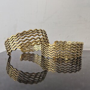 Op de afbeelding: Twee gouden metalen armbanden met een golvend ontwerp. De armbanden zijn op elkaar gestapeld.