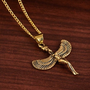 May include: Gold necklace with a pendant of a winged woman figure. The pendant is detailed with intricate lines and has a gold finish.