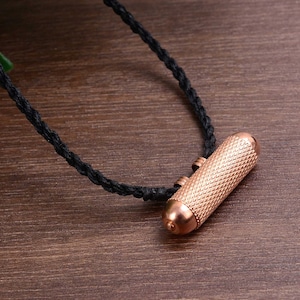 May include: A copper-colored cylindrical pendant with a textured surface hangs from a black cord. The pendant is a traditional Tibetan amulet.