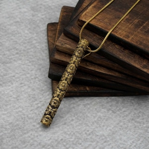 May include: A gold necklace with a long, cylindrical pendant featuring a floral design. The pendant is hanging on a stack of wooden coasters.