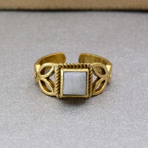 Op de afbeelding: Een gouden ring met een sierlijk ontwerp en een witte vierkante steen in het midden.