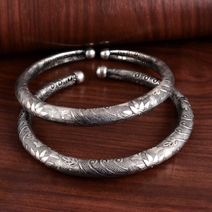 Puede incluir: Dos brazaletes de plata con intrincados diseños florales. Los brazaletes están apilados uno encima del otro y tienen un acabado brillante.