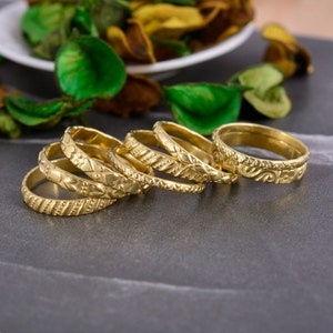 Puede incluir: Cinco anillos apilables de color dorado con intrincados diseños grabados. Los anillos están apilados sobre una superficie gris.