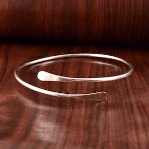 May include: A silver bangle bracelet with a simple, curved design. The bracelet is open and has a smooth, polished finish.