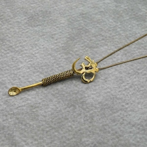 May include: A gold Om symbol pendant necklace with a small spoon attached to the bottom. The spoon is attached to the pendant with a twisted metal design.