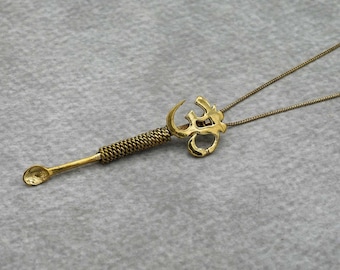 May include: A gold Om symbol pendant necklace with a small spoon attached to the bottom. The spoon is attached to the pendant with a twisted metal design.