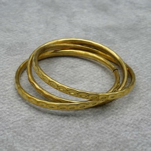 May include: Three gold-toned metal bangles with a textured, hammered design. The bangles are stacked on top of each other.