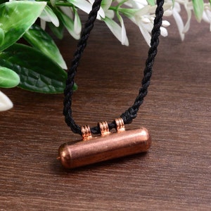 May include: A copper pendant with three decorative rings hangs from a black cord necklace. The pendant is cylindrical and has a smooth, polished finish.
