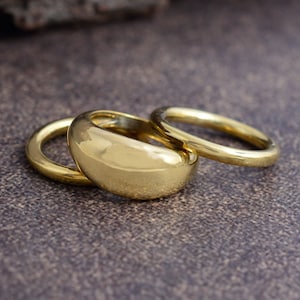 Könnte beinhalten: Drei goldene Ringe, einer mit abgerundeter, glatter Oberseite und zwei dünnere Bänder.