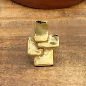May include: A gold-colored, abstract cross-shaped ring. The ring is made of multiple rectangular shapes that are joined together. The ring is sitting on a wooden surface.