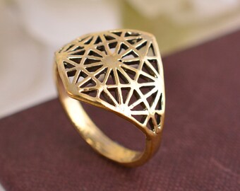 Puede incluir: Un anillo de color dorado con un diseño geométrico calado. El anillo tiene forma de diamante y presenta un patrón delicado e intrincado.