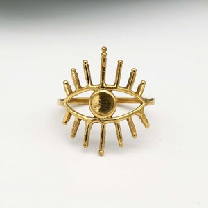 May include: A gold-colored ring with an eye design. The eye is outlined in a thin metal and has a round pupil. The eye is surrounded by a ring of thin metal rays.