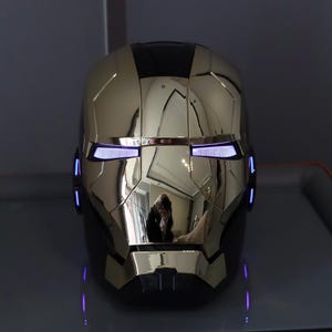 Könnte beinhalten: Ein gold-schwarzer Iron Man-Helm mit leuchtend blauen Augen und Akzenten. Der Helm ist hochreflektierend, mit einer polierten Oberfläche. Das Design basiert auf dem Marvel Comics-Superhelden.