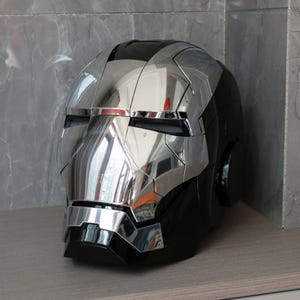 Può includere: Un casco argento e nero con finitura a specchio. Il casco ha un design dettagliato con una visiera nera e un cinturino per il mento nero.