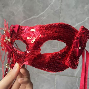 Op de afbeelding: Een rood, met pailletten versierd masker voor een gemaskerd bal met een strik aan elke kant en decoratieve veren. Het masker heeft ooggaten en wordt vastgehouden door een rood lint. Het masker is versierd met kleine gouden kralen.