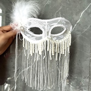 Könnte beinhalten: Weiße Spitzen-Maskenmaske mit Perlenfransen und Federakzent. Die Maske hat ein kompliziertes florales Spitzendesign und ist mit kleinen Perlen und einer zarten Kette verziert.