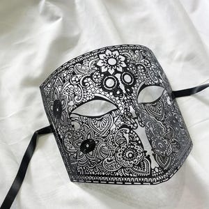 Puede incluir: Una máscara de carnaval en blanco y negro con intrincados diseños florales y de cachemira. La máscara tiene aberturas para los ojos y cintas negras. El diseño es simétrico, con adornos detallados en la superficie.