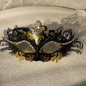 Puede incluir: Una máscara de carnaval negra y dorada con intrincados detalles de filigrana. La máscara presenta brillantes pedrería a lo largo de las aberturas de los ojos y una corona decorativa. Ideal para un evento formal o una fiesta de disfraces.