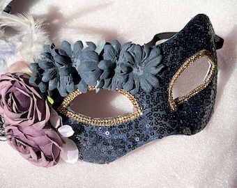 May include: A black masquerade mask with gold trim and a floral design. The mask is decorated with purple and white flowers and sequins.