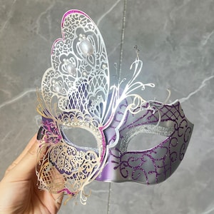 Pode incluir: Uma máscara de baile decorativa com um design de asa de borboleta. A máscara apresenta intrincados filigranas prateados, glitter brilhante e detalhes em roxo e prata. A máscara é adornada com strass e pérolas.
