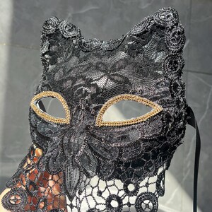 以下が含まれることがあります： 黒いレースのキャットアイマスク。目の周りに金色のトリムが付いています。マスクは繊細な花柄で、サテンのリボンタイが付いています。マスクは目と鼻を覆うように設計されています。