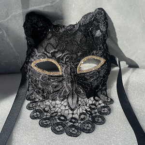 以下が含まれることがあります： 黒いレースの猫マスク。目の周りに金色のトリムが付いています。マスクは装飾的なレースのデザインで、黒いサテンのリボンが付いています。マスクは目と顔の上部を覆うように設計されています。