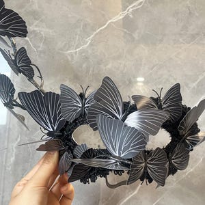 May include: A black masquerade mask decorated with multiple butterfly features. The butterflies have white vein detailing on their wings. The mask is set against a grey marble background, highlighting the intricate design and craftsmanship of the accessory.