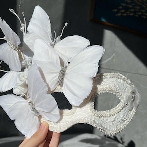 May include: A white lace masquerade mask adorned with delicate white feather butterflies and sparkling embellishments. The mask features intricate lace detailing and is accented with small white flowers and pearls, creating an elegant accessory.