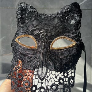 以下が含まれることがあります： 黒いレースの猫マスク、金色の縁取りの目の開口部。 マスクは繊細なレースのデザイン、猫の耳の形をしており、目の周りにキラキラ光るラインストーンが飾られています。 マスクを固定するための黒いリボンが付いています。