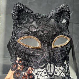 以下が含まれることがあります： 黒いレースのキャットアイマスク。目の周りに金色のトリムが付いています。マスクは繊細な花のレース模様で、猫の耳が付いています。マスクを固定するための黒いリボンが付いています。