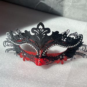 May include: Black and red masquerade mask with intricate filigree design. The mask features sparkling rhinestones and a glittery red finish on the lower half. A decorative accessory for parties and events.