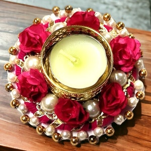 Könnte beinhalten: Ein dekorativer Kerzenhalter mit einer brennenden Kerze in einem goldfarbenen Metallrahmen. Er ist mit roten Stoffblumen, Kunstperlen und goldfarbenen Perlen verziert. Der runde Kerzenhalter steht auf einer Holzoberfläche.